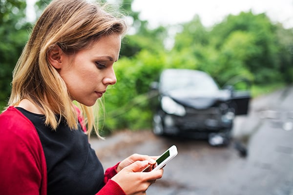a-young-woman-with-smartphone-by-the-damaged-car-7FPTKGR